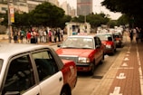 Several red and white taxis are queued up along a city street with a group of people waiting on the sidewalk nearby. Tall buildings and trees border the scene, creating a typical urban environment.
