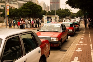 Several red and white taxis are queued up along a city street with a group of people waiting on the sidewalk nearby. Tall buildings and trees border the scene, creating a typical urban environment.