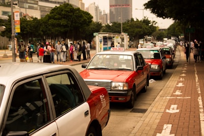 Several red and white taxis are queued up along a city street with a group of people waiting on the sidewalk nearby. Tall buildings and trees border the scene, creating a typical urban environment.