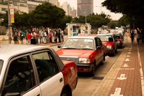 Several red and white taxis are queued up along a city street with a group of people waiting on the sidewalk nearby. Tall buildings and trees border the scene, creating a typical urban environment.