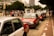 Several red and white taxis are queued up along a city street with a group of people waiting on the sidewalk nearby. Tall buildings and trees border the scene, creating a typical urban environment.
