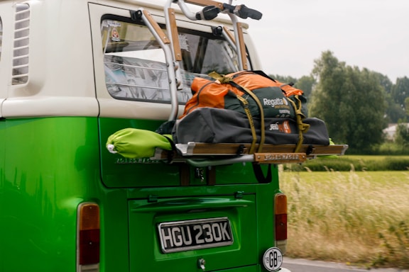 A vintage green and white van with a luggage rack on the back is loaded with an orange and gray duffel bag. The bag is secured with straps. In the background, there is a blurred view of greenery and trees indicating motion.