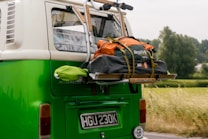 A vintage green and white van with a luggage rack on the back is loaded with an orange and gray duffel bag. The bag is secured with straps. In the background, there is a blurred view of greenery and trees indicating motion.