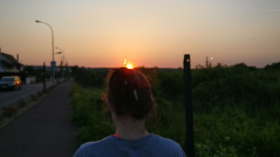 A person with tied hair is seen from behind, walking along a path bordered by greenery. The scene captures a vibrant sunset on the horizon, with streetlights lining the road on the left side. A car is visible on the roadway, and the sky transitions from pink to orange and deep blue.