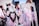 Elegant white chairs are adorned with decorative elements, including soft pink tulle, fabric roses, and woven heart-shaped ornaments. The background is blurred, suggesting an outdoor setting with people dressed formally.