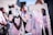 Elegant white chairs are adorned with decorative elements, including soft pink tulle, fabric roses, and woven heart-shaped ornaments. The background is blurred, suggesting an outdoor setting with people dressed formally.