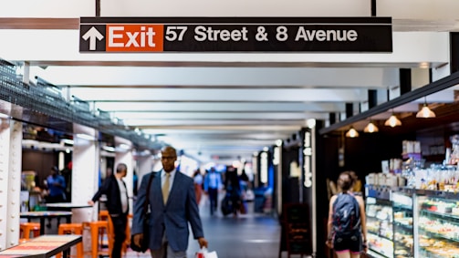 A busy indoor corridor featuring a large exit sign that directs to 57 Street and 8 Avenue. The area is well-lit, with several people walking and engaging with vendors on the sides. A man in a suit is prominent in the foreground, and food displays are visible on the right.