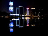 Nighttime cityscape with skyscrapers illuminated in electric blue and gold accents reflecting on a river.