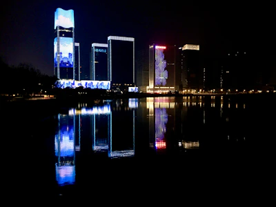 Nighttime cityscape with skyscrapers illuminated in electric blue and gold accents reflecting on a river.