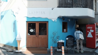 A quaint storefront with blue and white exterior, featuring a wooden door with an 'open' sign. An oversized ice cream cone model is placed by the entrance. There is a red vending machine with a polar bear design on the right side. Two people are standing near a menu board outside the shop. The word 'Laitier' is emblazoned above the door.