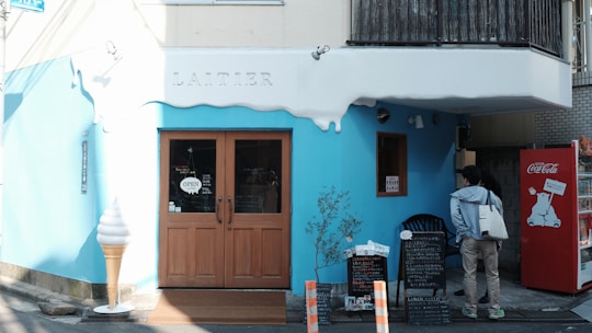 A quaint storefront with blue and white exterior, featuring a wooden door with an 'open' sign. An oversized ice cream cone model is placed by the entrance. There is a red vending machine with a polar bear design on the right side. Two people are standing near a menu board outside the shop. The word 'Laitier' is emblazoned above the door.