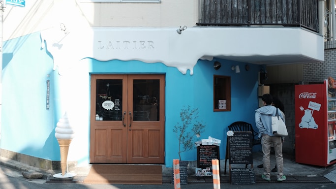 A quaint storefront with blue and white exterior, featuring a wooden door with an 'open' sign. An oversized ice cream cone model is placed by the entrance. There is a red vending machine with a polar bear design on the right side. Two people are standing near a menu board outside the shop. The word 'Laitier' is emblazoned above the door.