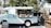 A vintage food truck painted light blue and white is parked outdoors. The truck is set up for business with an open service window displaying various items like baskets and condiments. A woman stands beside the truck wearing an apron. The surrounding area includes a small collection of wooden furniture and decorative items, likely indicating a market or outdoor gathering. Large trees and a building in the background suggest an urban setting.