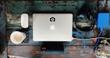 On a rustic wooden surface, a silver laptop with an Apple logo occupies the center, accompanied by a white wireless mouse on the left. A steaming cup of coffee sits on a patterned coaster nearby. To the right, black headphones rest beside a blue external hard drive connected to the laptop. Below, a black portable speaker is positioned horizontally.