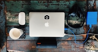 On a rustic wooden surface, a silver laptop with an Apple logo occupies the center, accompanied by a white wireless mouse on the left. A steaming cup of coffee sits on a patterned coaster nearby. To the right, black headphones rest beside a blue external hard drive connected to the laptop. Below, a black portable speaker is positioned horizontally.