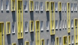 A building facade with a series of windows arranged in a repetitive pattern. The exterior is gray with bright yellow vertical accents surrounding the windows, creating a modern and geometric look. Shadows from the window frames add depth to the flat surface.