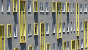 A building facade with a series of windows arranged in a repetitive pattern. The exterior is gray with bright yellow vertical accents surrounding the windows, creating a modern and geometric look. Shadows from the window frames add depth to the flat surface.