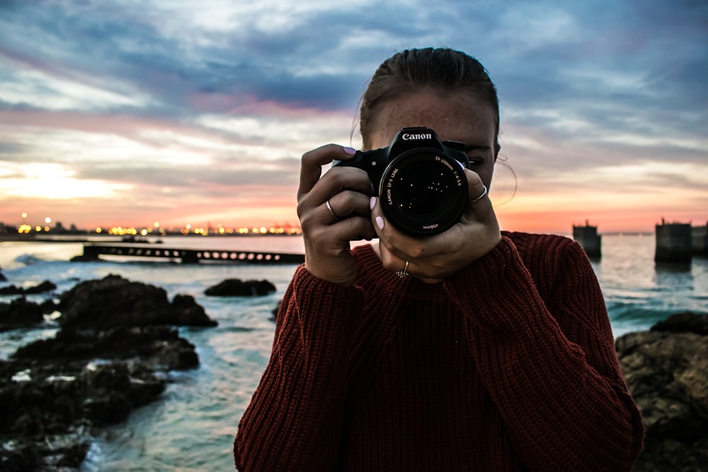 foto di donna che tiene in mano una fotocamera reflex digitale Canon nera vicino allo specchio d'acqua