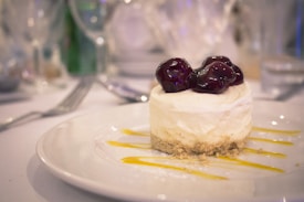 A dessert featuring a small cheesecake topped with glossy cherries sits elegantly on a white plate. The dessert is garnished with streaks of yellow sauce, and the background includes softly blurred glassware and table settings, suggesting a formal dining setting.