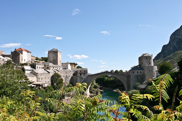 A historic stone bridge arches gracefully over a turquoise river, flanked by traditional stone buildings with various geometric shapes and red-tiled roofs. Lush greenery frames the foreground, while a rugged mountain rises in the background under a clear blue sky.