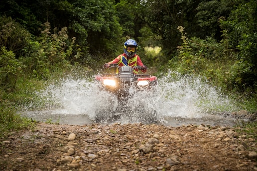 A person wearing a helmet and protective gear is riding an all-terrain vehicle (ATV) through a shallow water stream in a dense, green forest. The vehicle is splashing water up as it moves forward, creating a dynamic action scene. The path is rugged and lined with rocks.