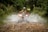 A person wearing a helmet and protective gear is riding an all-terrain vehicle (ATV) through a shallow water stream in a dense, green forest. The vehicle is splashing water up as it moves forward, creating a dynamic action scene. The path is rugged and lined with rocks.