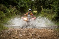 A person wearing a helmet and protective gear is riding an all-terrain vehicle (ATV) through a shallow water stream in a dense, green forest. The vehicle is splashing water up as it moves forward, creating a dynamic action scene. The path is rugged and lined with rocks.
