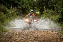 A person wearing a helmet and protective gear is riding an all-terrain vehicle (ATV) through a shallow water stream in a dense, green forest. The vehicle is splashing water up as it moves forward, creating a dynamic action scene. The path is rugged and lined with rocks.