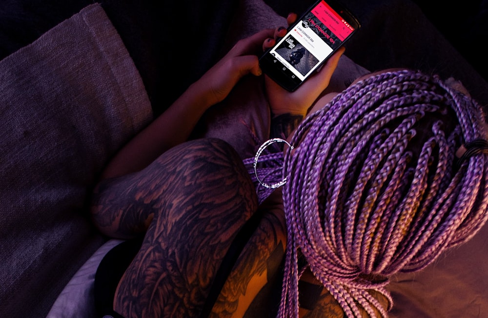 A woman in purple dreadlocks with tattoos on her back looking at a music application on a phone