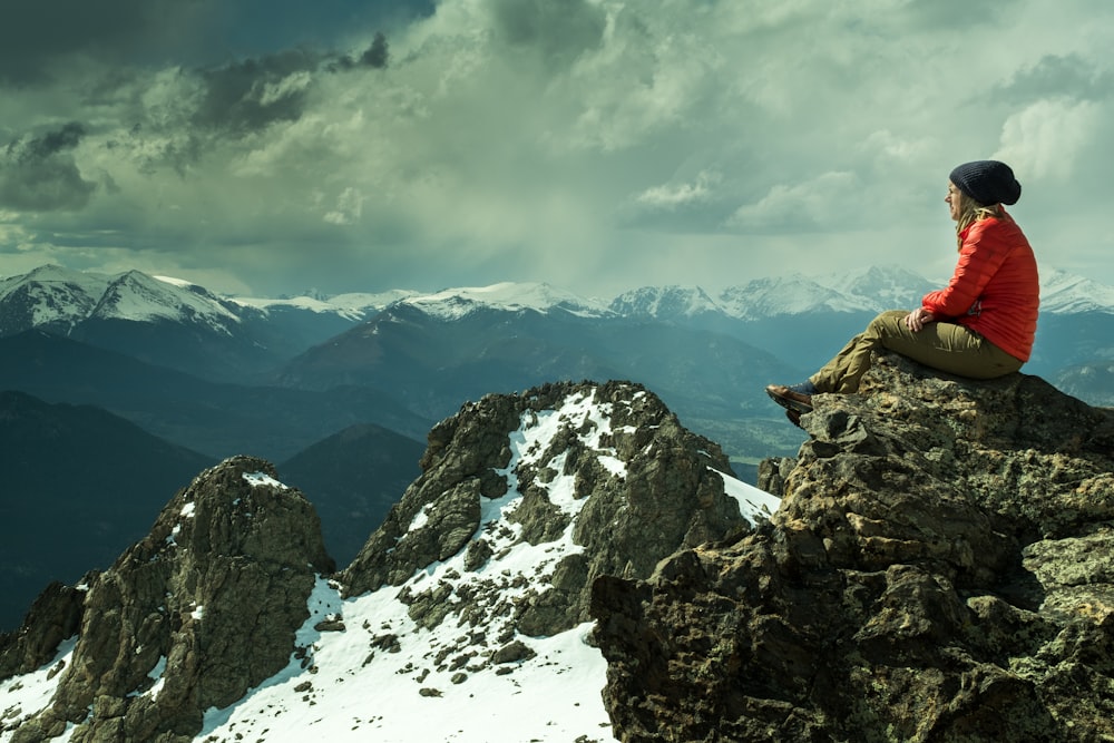 Person, die auf einem Felsen über einem schneebedeckten Berg unter bewölktem Himmel sitzt