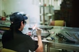 A person in a workshop is using a blowtorch to work on a small object. They are wearing a cap, glasses, and a glove for protection. The workspace consists of a metal table with various tools and materials scattered around, including glass rods. The scene has a casual and focused atmosphere.