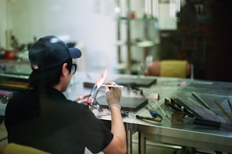 A person in a workshop is using a blowtorch to work on a small object. They are wearing a cap, glasses, and a glove for protection. The workspace consists of a metal table with various tools and materials scattered around, including glass rods. The scene has a casual and focused atmosphere.