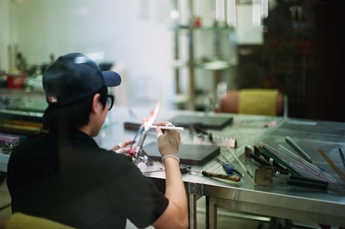 A person in a workshop is using a blowtorch to work on a small object. They are wearing a cap, glasses, and a glove for protection. The workspace consists of a metal table with various tools and materials scattered around, including glass rods. The scene has a casual and focused atmosphere.