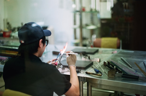 A person in a workshop is using a blowtorch to work on a small object. They are wearing a cap, glasses, and a glove for protection. The workspace consists of a metal table with various tools and materials scattered around, including glass rods. The scene has a casual and focused atmosphere.