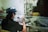 A person in a workshop is using a blowtorch to work on a small object. They are wearing a cap, glasses, and a glove for protection. The workspace consists of a metal table with various tools and materials scattered around, including glass rods. The scene has a casual and focused atmosphere.