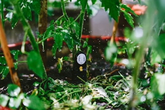 A No-Face character figurine is nestled in a lush garden setting among vibrant green leaves and stems of plants. Water droplets are visibly hanging from the leaves, suggesting recent rainfall or watering.