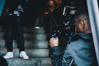A person operating a professional video camera is filming someone who is standing on a staircase. The stairway is made of concrete, and the lighting is dim, suggesting an indoor setting. The individual being filmed is blurred and partially visible, particularly notable are their white sneakers.