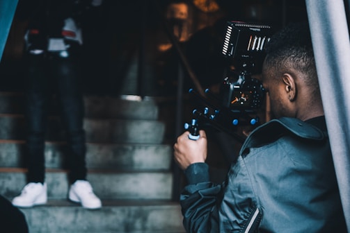 A person operating a professional video camera is filming someone who is standing on a staircase. The stairway is made of concrete, and the lighting is dim, suggesting an indoor setting. The individual being filmed is blurred and partially visible, particularly notable are their white sneakers.