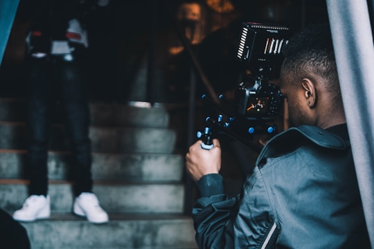 A person operating a professional video camera is filming someone who is standing on a staircase. The stairway is made of concrete, and the lighting is dim, suggesting an indoor setting. The individual being filmed is blurred and partially visible, particularly notable are their white sneakers.