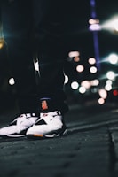 A close-up view of a person wearing stylish sneakers, standing on a street at night. The image is focused on the shoes, with blurred city lights creating an atmospheric bokeh effect in the background.