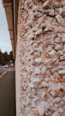 A close-up view of a textured wall composed of small, rounded stones set into concrete. The stones range from white to light brown, creating a speckled pattern. In the background, a road with cars is visible, leading to a distant set of traffic lights. Trees line the side of the road under a cloudy sky.