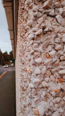 A close-up view of a textured wall composed of small, rounded stones set into concrete. The stones range from white to light brown, creating a speckled pattern. In the background, a road with cars is visible, leading to a distant set of traffic lights. Trees line the side of the road under a cloudy sky.