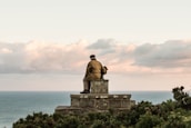 An elderly person wearing a brown coat and cap sits on a stone bench overlooking a vast ocean. The distant horizon line separates the calm seawater from a sky filled with soft, scattered clouds. The scene is surrounded by some green bushes.