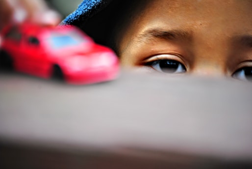 A close-up of a child's eyes peering over a surface with a blurred red toy car in the foreground. The focus is on the child's intent gaze, suggesting curiosity or concentration.