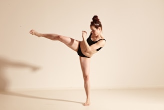 A person wearing a black outfit is performing a high kick against a plain, light-colored background. The pose suggests energy and balance, with a focused facial expression and tension in the raised leg. The individual has their hair tied up, emphasizing a sense of discipline and strength.