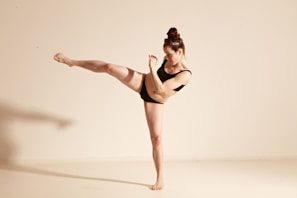 A person wearing a black outfit is performing a high kick against a plain, light-colored background. The pose suggests energy and balance, with a focused facial expression and tension in the raised leg. The individual has their hair tied up, emphasizing a sense of discipline and strength.