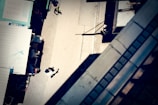 An aerial view of a street scene with a few people walking. The image includes market stalls on the left side, casting shadows onto the pavement. A person in a white shirt is visible walking along the sidewalk. Architectural elements on the right create geometric patterns.