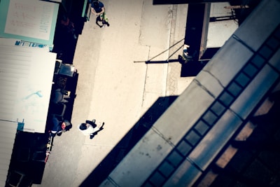 An aerial view of a street scene with a few people walking. The image includes market stalls on the left side, casting shadows onto the pavement. A person in a white shirt is visible walking along the sidewalk. Architectural elements on the right create geometric patterns.