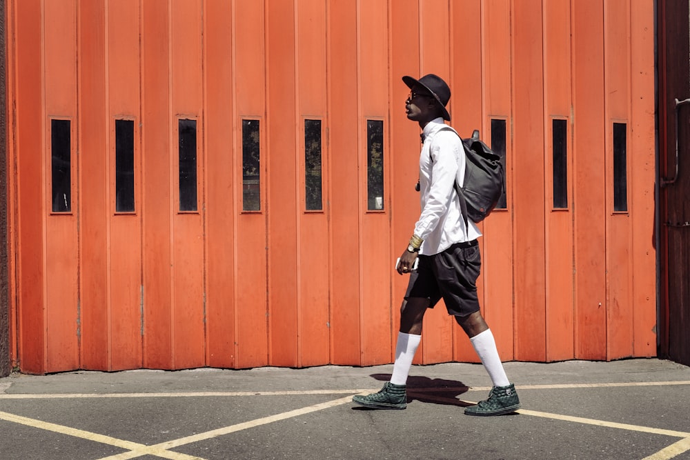 man wearing black hat and white dress shirt walking on street near orange gate