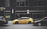 A yellow taxi is parked on the street between two black cars in front of a large building with dark windows. The building has signage for the Hard Rock Hotel and another sign advertising Jamaican cuisine. A vertical light fixture decorates the facade, and there are no visible reflections in the windows.